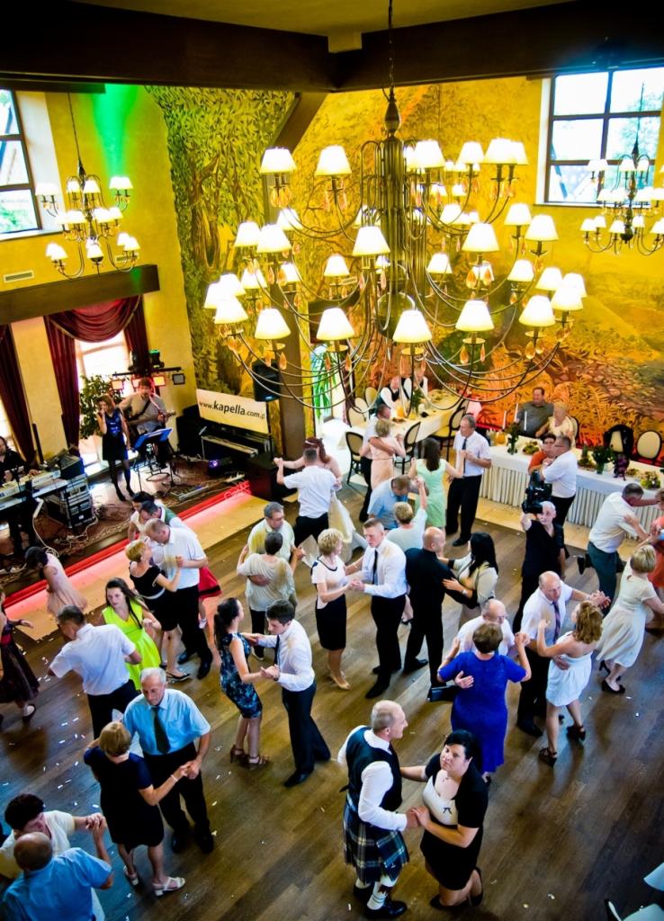 Sala balowa z parkietem wypełniona tańczącymi parami, w tle zespół muzyczny grający na scenie podświetlonej zielonym światłem, nad głowami gości bogato zdobiony żyrandol, na ścianie malowidło...