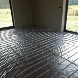 Rozłożona instalacja ogrzewania podłogowego na folii termoizolacyjnej w surowym wnętrzu z dużymi oknami, widok na zewnątrz.