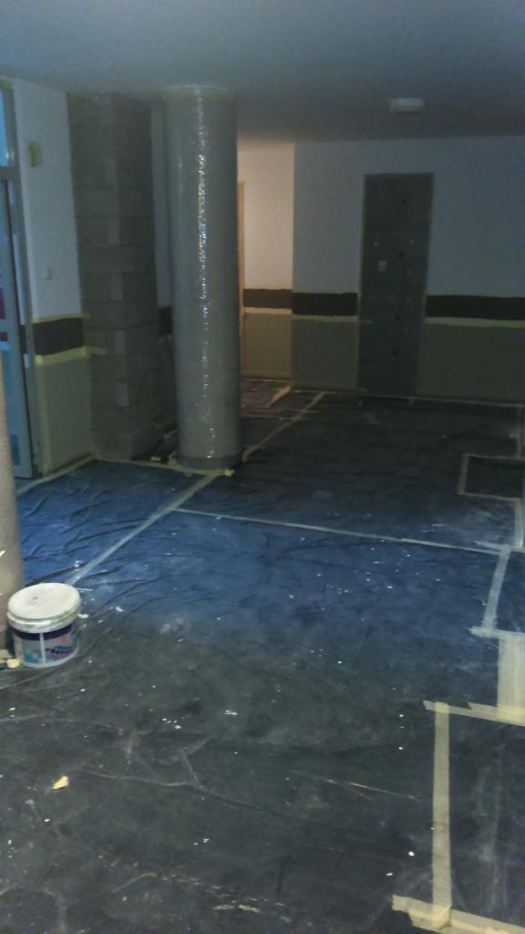 Przygotowania do malowania biura: podłoga zabezpieczona folią, kolumny owinięte, ściany z taśmą malarską, widoczne puszki z farbą.