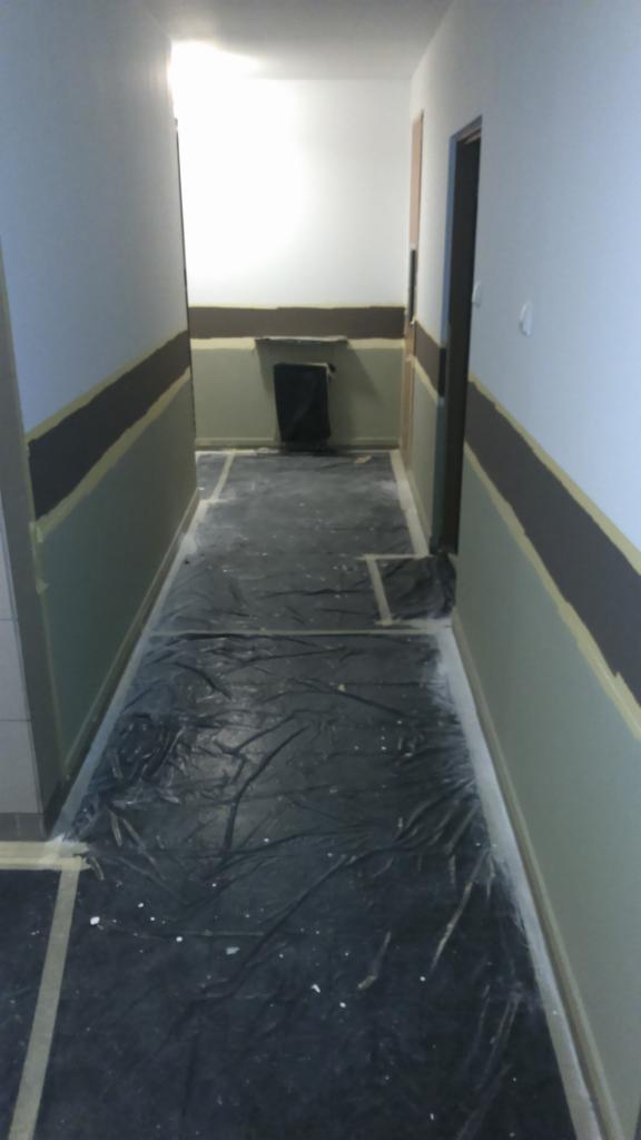 Przygotowanie do malowania biura w Warszawie: podłoga zabezpieczona folią, ściany z taśmą malarską i pomalowanym pasem w kolorze czekoladowym.