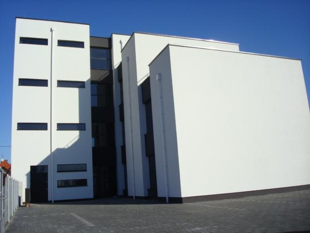 Nowoczesny, biały budynek wielorodzinny z ciemnymi oknami i elementami wykończeniowymi, sfotografowany w słoneczny dzień z niskiej perspektywy.