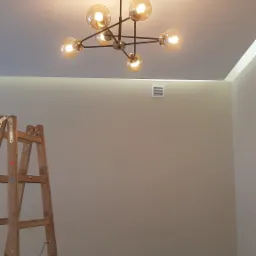 Nowoczesny żyrandol z sześcioma szklanymi kulami zamontowany na suficie, widoczna drewniana drabina oparta o ścianę, w tle kratka wentylacyjna i gniazdka elektryczne.