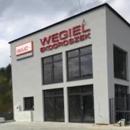 Budynek z dużym napisem 'WĘGIEL EKOGROSZEK' w kolorze czerwonym, widoczne okna i logo firmy GLUC na ścianie, szary budynek z płaskim dachem, ogrodzenie z metalowych prętów.