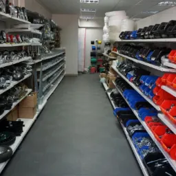 Wnętrze sklepu z szerokim asortymentem kół, kółek i akcesoriów montażowych, ułożonych w rzędach na białych półkach, z widocznymi pojemnikami w kolorach czerwonym, niebieskim i czarnym.