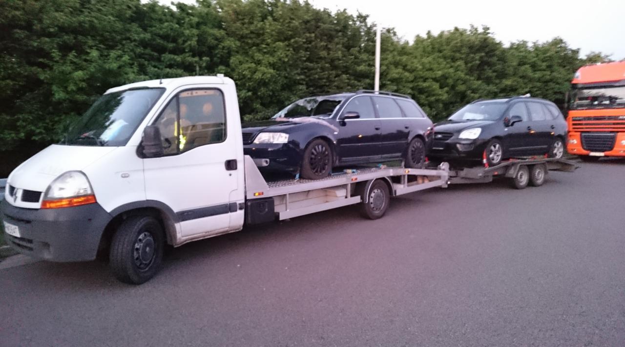 Biała laweta Renault z dwoma ciemnymi samochodami osobowymi na platformie i przyczepie; w tle pomarańczowa ciężarówka i zieleń drzew.