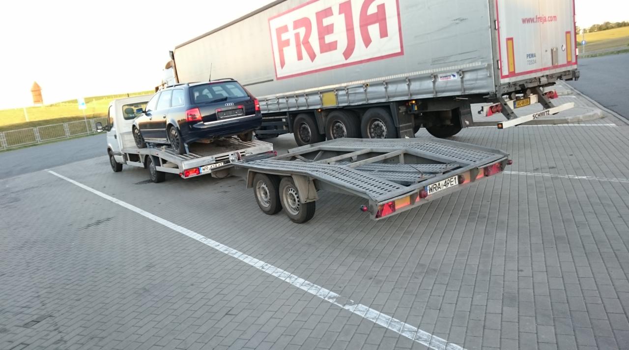 Laweta z Audi A6 na platformie, obok zaparkowana ciężarówka Freja. Transport samochodów realizowany na parkingu z kostki brukowej.