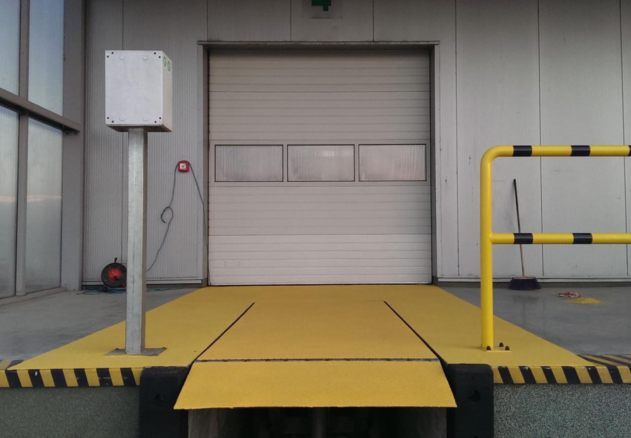 Rampa załadunkowa z żółtą, antypoślizgową nawierzchnią przed bramą przemysłową, oznaczona czarno-żółtymi pasami bezpieczeństwa, z widoczną barierą ochronną i elementami infrastruktury magazynowej.