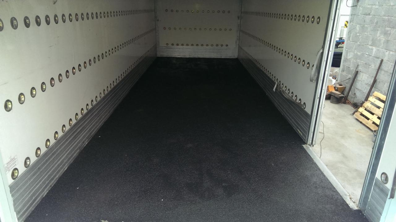 Wnętrze naczepy z nową, czarną, antypoślizgową powierzchnią podłogi, widoczne mocowania na ścianach i fragment otoczenia zewnętrznego z paletami.