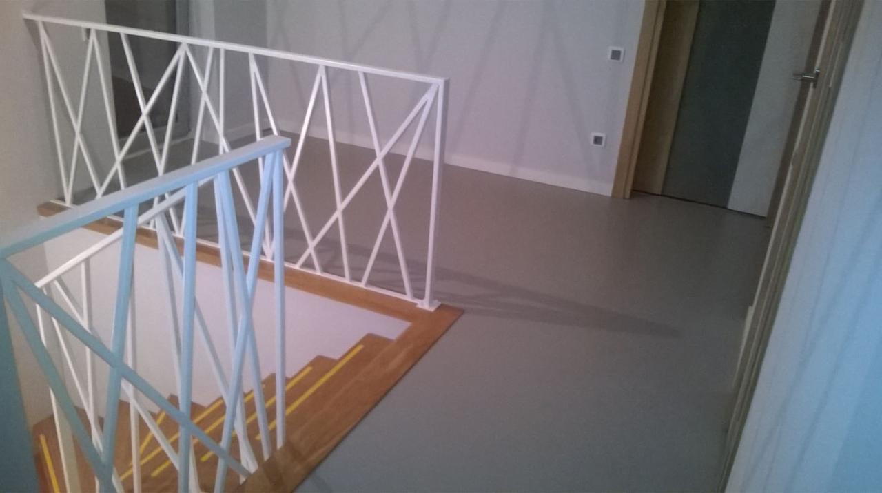 Szara, gładka podłoga w korytarzu z widocznymi schodami z drewnianymi stopniami zabezpieczonymi żółtymi paskami antypoślizgowymi i białymi, nowoczesnymi barierkami ze skośnymi elementami.