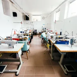 Jasne wnętrze szwalni z rzędem stanowisk pracy wyposażonych w maszyny do szycia marki Siruba, oświetlenie punktowe i akcesoria krawieckie.