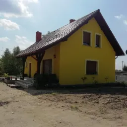 Żółty dom jednorodzinny z brązowym dachem i stolarką okienną, widok z boku na tle błękitnego nieba z chmurami, teren wokół domu z ziemią po pracach budowlanych.