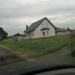 Nowy dom jednorodzinny z jasną elewacją i ciemnym dachem, widziany z perspektywy kierowcy samochodu na tle pochmurnego nieba.