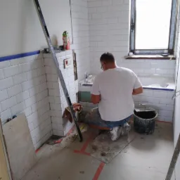 Prace wykończeniowe w łazience: mężczyzna w białej koszulce układa białe płytki na ścianie, widoczna poziomica, wiadro i fragment wanny.