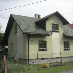 Jasnozielony dom jednorodzinny z ciemnozielonym dachem z blachodachówki, widok z zewnątrz, z numerem 261 na elewacji i kwiatami w oknach.