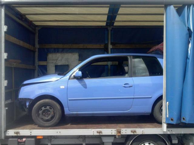 Niebieski samochód osobowy bez przedniego zderzaka, transportowany wewnątrz busa z plandeką. Widok z boku, oświetlenie dzienne.