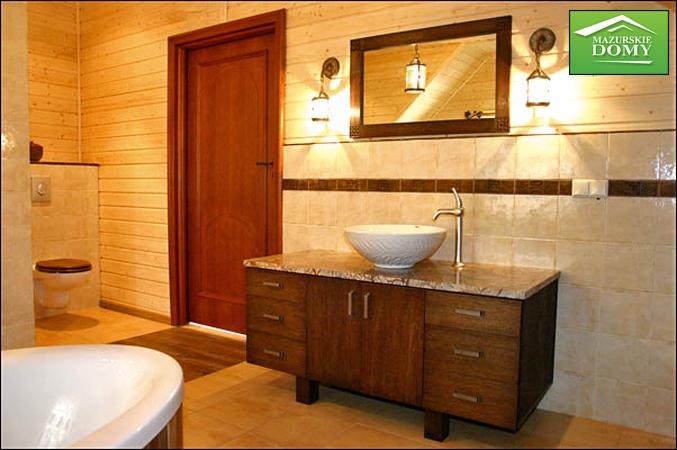 Rustykalna łazienka w domu z bali: drewniane ściany, szafka z umywalką, lustro z oświetleniem. Widoczny fragment wanny i toalety. Ciepłe oświetlenie, przytulny klimat.