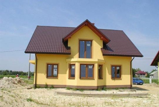 Żółty dom jednorodzinny z brązowym dachem, widoczny z przodu, z niezagospodarowanym terenem wokół. Okna z brązowymi ramami. Błękitne niebo w tle.