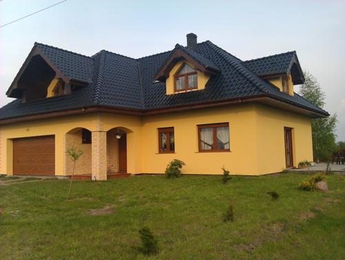 Żółty dom jednorodzinny z ciemnym dachem, widok z trawnika. Jasna elewacja, drewniane okna i brama garażowa. Uwagę zwraca wykończenie kolumn wejściowych.