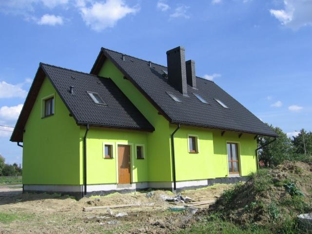 Nowo pomalowany dom w odcieniach zieleni z ciemnym dachem i dwoma kominami, otoczony ziemią i zielenią, pod błękitnym niebem z lekkimi chmurami.