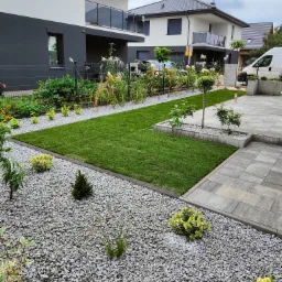 Nowoczesny ogród z trawnikiem z rolki, rabatami z kamienia i ozdobnymi krzewami na tle domu z balkonem. Widoczny fragment podjazdu z kostki.