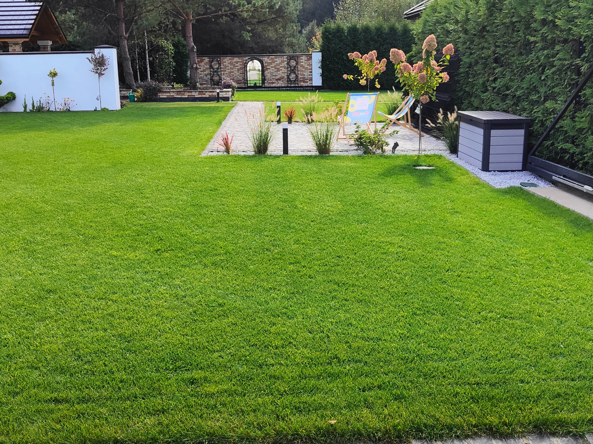 Zadbany trawnik z kostką brukową, dekoracyjnymi trawami i elementami architektury ogrodowej, takimi jak biały mur i pergola z cegły.