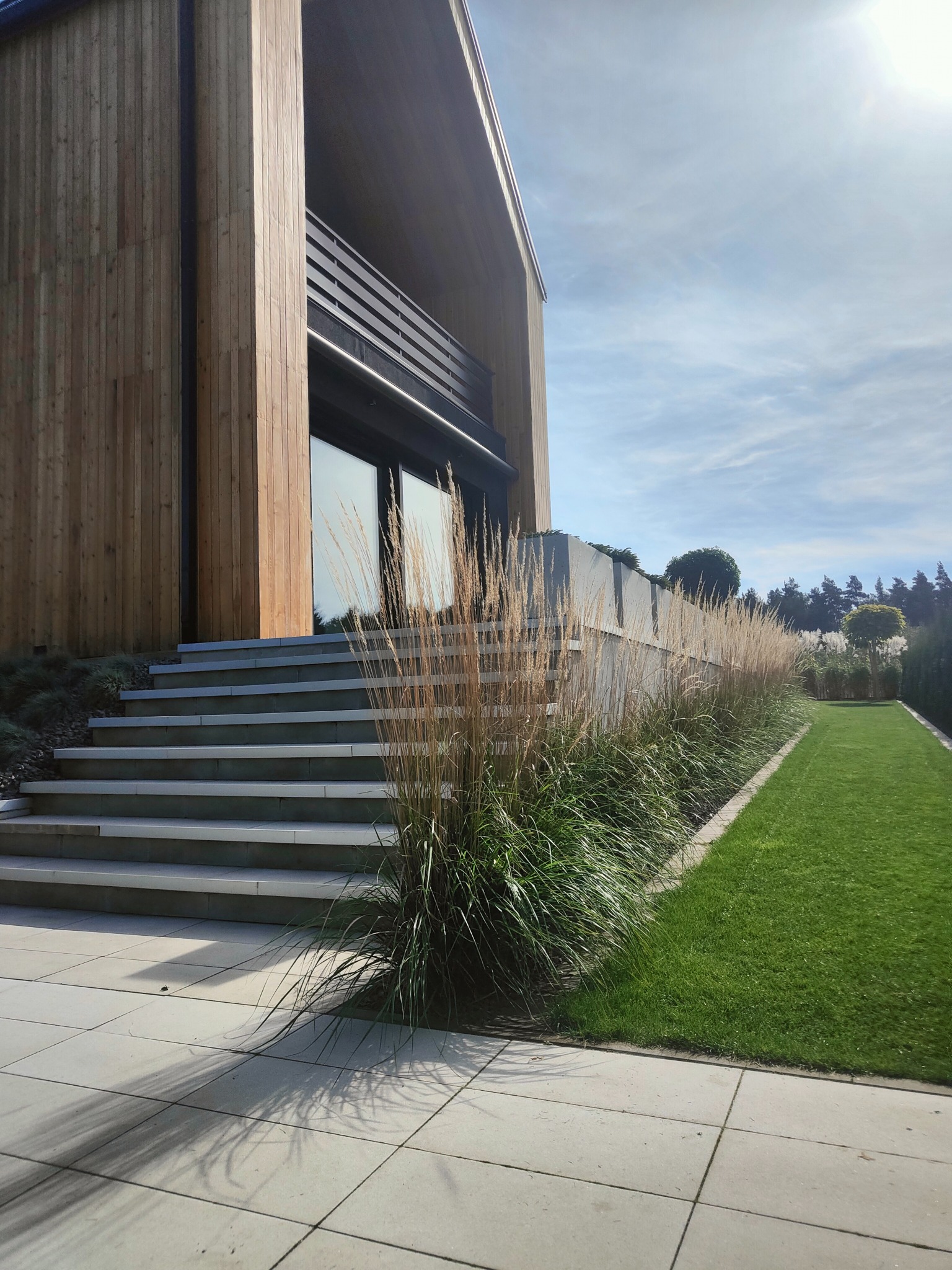 Nowoczesny ogród z trawą ozdobną i równo przystrzyżonym trawnikiem, prowadzący do drewnianego domu. Kąt ujęcia podkreśla harmonię architektury i krajobrazu.