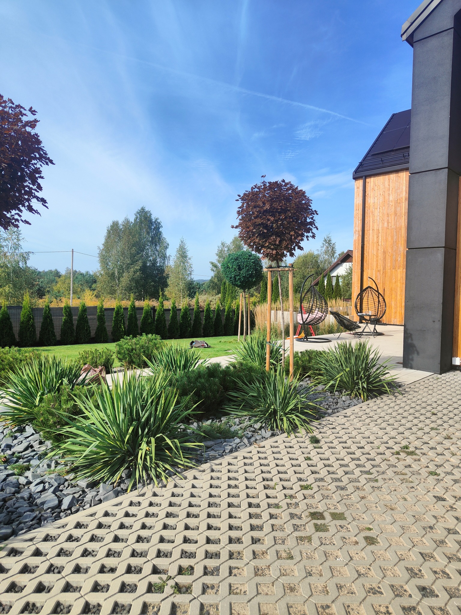 Zadbany ogród z ozdobnymi krzewami i drzewami, nowoczesny dom z drewnianą elewacją, dekoracyjne kamienie i ażurowe płyty chodnikowe. Dwie wiszące huśtawki.