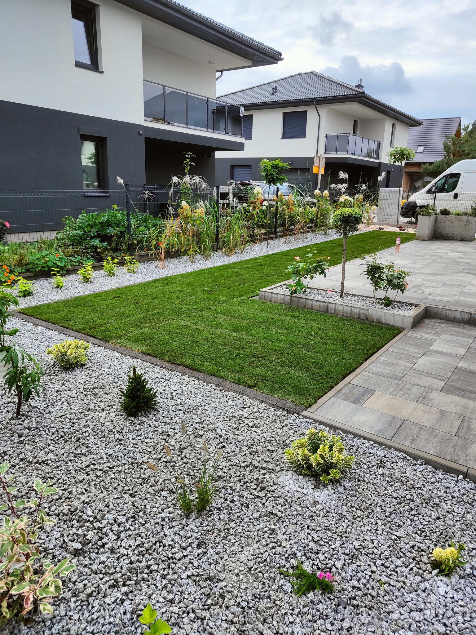 Nowoczesny ogród z trawnikiem z rolki, rabatami z kamienia i ozdobnymi krzewami na tle domu z balkonem. Widoczny fragment podjazdu z kostki.