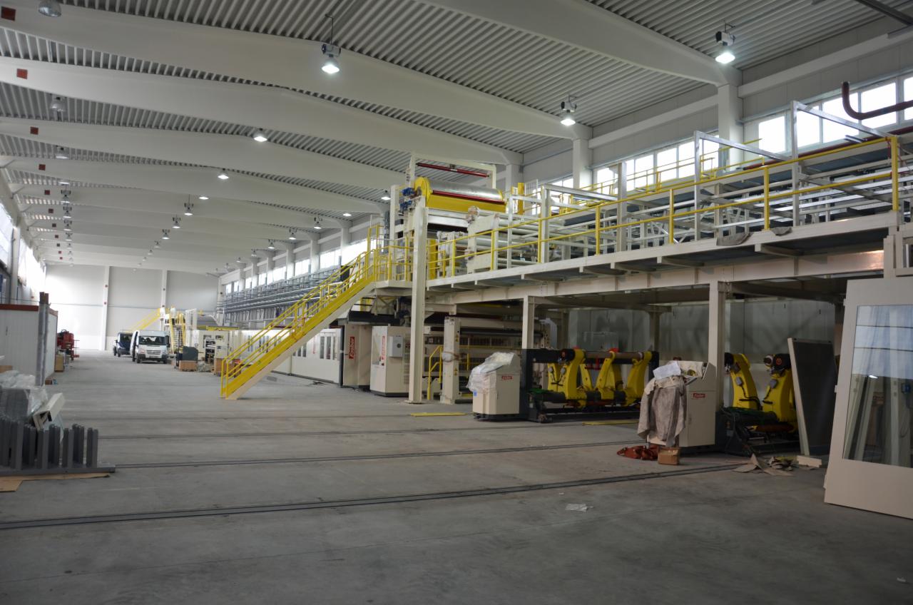 Rozległa hala produkcyjna z linią technologiczną do obróbki materiałów, widoczne żółte schody i platformy obsługowe, oświetlenie jarzeniowe pod sufitem.