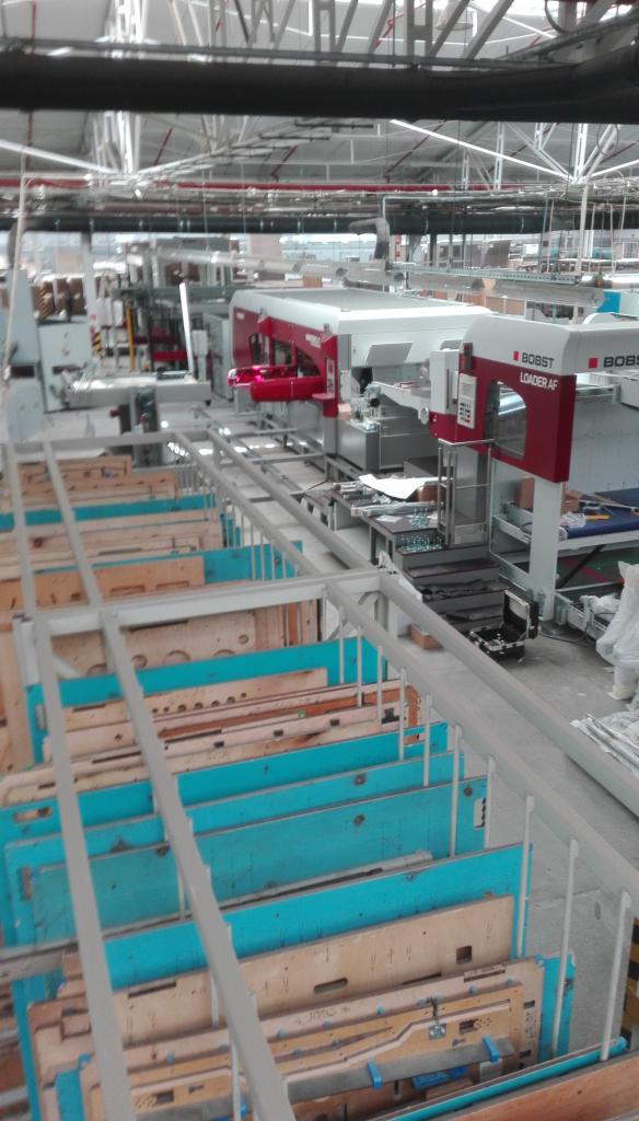 Widok z góry na linię produkcyjną z maszyną Bobst Loader AF, z widocznymi elementami do obróbki w turkusowo-drewnianych pojemnikach, w hali fabrycznej.