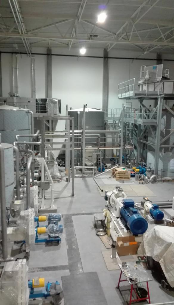 Wnętrze hali produkcyjnej z zainstalowanymi maszynami i zbiornikami, widoczne rury, silniki oraz elementy konstrukcyjne, częściowo jeszcze zabezpieczone folią, oświetlenie górne.