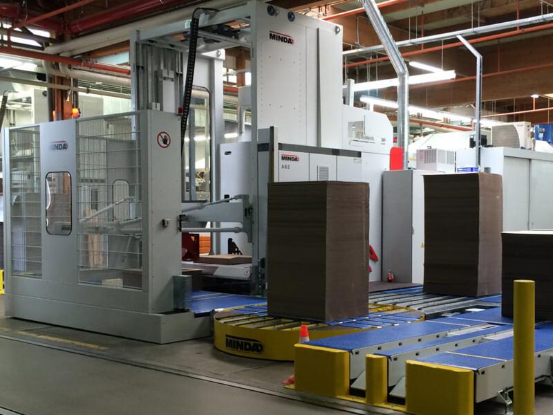 Duża, automatyczna maszyna przemysłowa marki MINDA transportująca stosy brązowych kartonów na niebieskim przenośniku taśmowym w hali produkcyjnej.