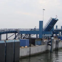 Widok na nabrzeże z rampą załadunkową i elementami konstrukcji stalowej, częściowo nad wodą. Widoczny dźwig i kontenery. Pochmurne niebo.