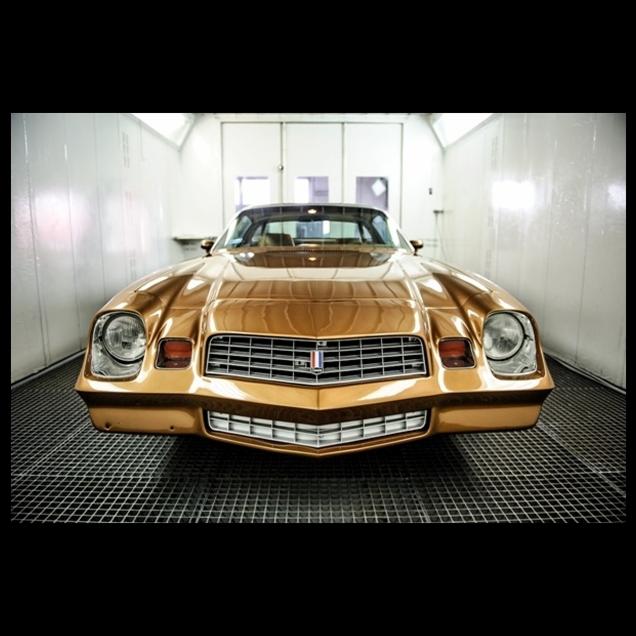 Złoty, odrestaurowany Chevrolet Camaro z 1978 roku, zaparkowany w jasnym pomieszczeniu z kratowaną podłogą. Widok z przodu, detale grilla i reflektorów.