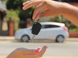 Dłoń wręcza kluczyk do białego, rozmytego auta w tle. Kluczyk ma czarny brelok. Druga dłoń z różowym manicure odbiera kluczyk. Słoneczny dzień.