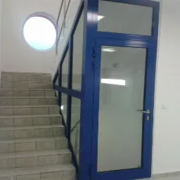 Klatka schodowa z niebieską, przeszkloną balustradą i drzwiami. Okrągłe okno na ścianie. Jasne płytki na schodach. Ujęcie z dołu schodów.