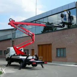 Czerwony podnośnik koszowy na białej ciężarówce, z dwoma pracownikami w koszu na tle budynku z dużymi oknami, podczas prac na wysokości.