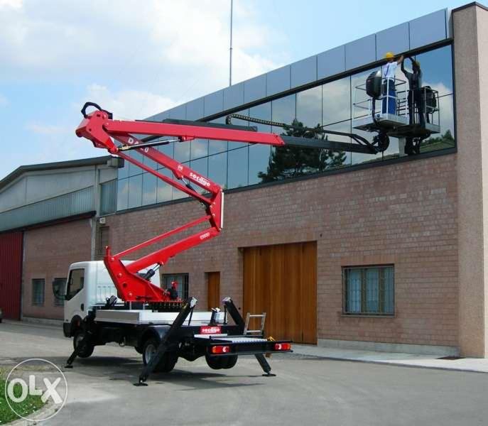 Czerwony podnośnik koszowy na białej ciężarówce, z dwoma pracownikami w koszu na tle budynku z dużymi oknami, podczas prac na wysokości.