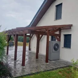 Drewniana pergola przy domu w Somoninie, z widoczną pompą ciepła i kostką brukową. Ujęcie pokazuje detale konstrukcji w pochmurny dzień.