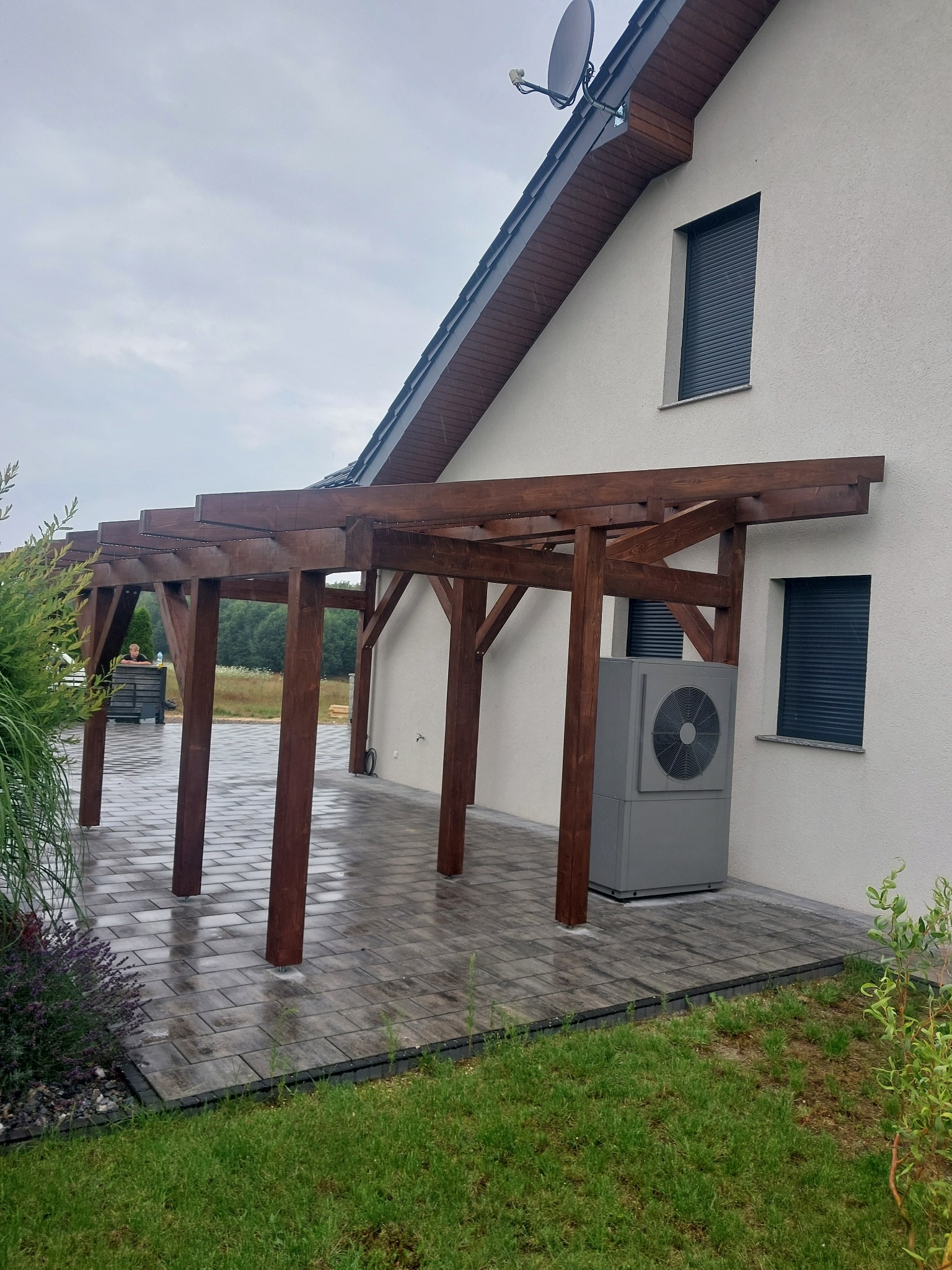 Drewniana pergola przy domu w Somoninie, z widoczną pompą ciepła i kostką brukową. Ujęcie pokazuje detale konstrukcji w pochmurny dzień.
