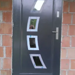 Nowoczesne, grafitowe drzwi wejściowe z czterema, ukośnymi szybkami, zamontowane w surowym murze z cegły. Widoczne ślady pianki montażowej.