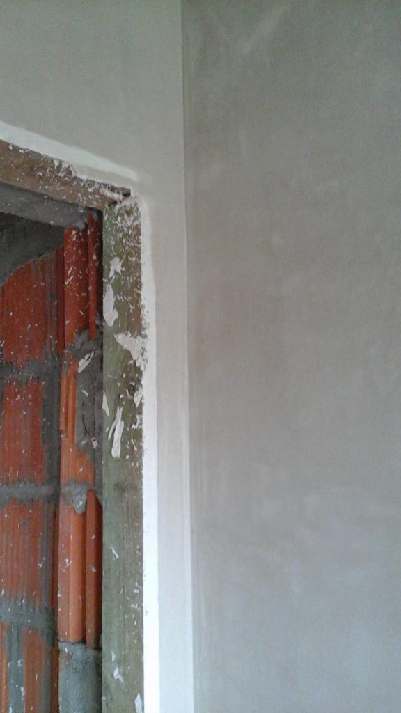 Ściana po nałożeniu tynku maszynowego, widoczna ościeżnica drzwiowa z cegły. Tynk świeży, jasny, gładki. Widoczne ślady po pracach wykończeniowych.