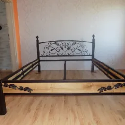 Metalowa rama łóżka z ozdobnym zagłówkiem na drewnianej podłodze. Elementy dekoracyjne w stylu retro. W tle ściana z subtelnym wzorem.