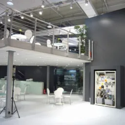 Wnętrze hali z antresolą ze stalową balustradą. Widoczne białe krzesła i stoliki, ekspozycja w zabudowie oraz oświetlenie sufitowe.