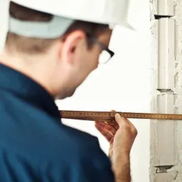 Mężczyzna w kasku mierzy otwór w ścianie z pustaków za pomocą drewnianej miarki, skupiony na dokładnym pomiarze, z widocznymi detalami struktury ściany.