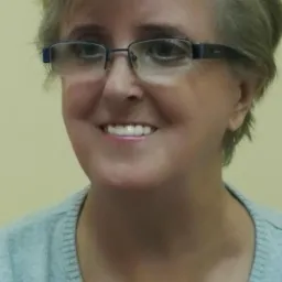 Portret uśmiechniętej kobiety w okularach o niebieskich oprawkach, z krótkimi blond włosami, na rozmytym żółtym tle.