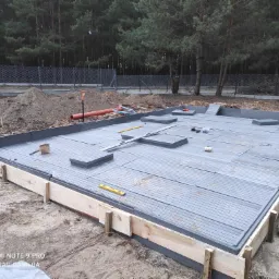 Przygotowanie podłoża pod budowę basenu z drewnianą ramą, widoczne deski podłogowe, poziomice i narzędzia na tle ogrodzenia z siatki i drzew