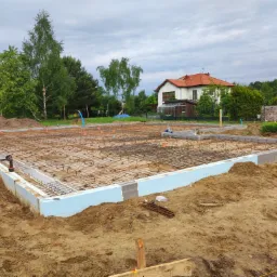 Zbrojenie fundamentów z użyciem siatki stalowej i szalunku z izolacją termiczną, widoczny pracownik przywiązujący druty, w tle dom jednorodzinny i zieleń.