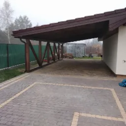 Ułożona kostka brukowa pod drewnianą wiatą przylegającą do budynku, widoczny fragment zielonego ogrodzenia i zarys drzew we mgle.