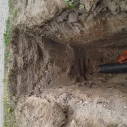 Wykop z rurą i taśmą ostrzegawczą. Ziemia, trawa i kostka brukowa w tle. Widok z góry.